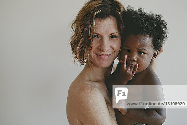 Porträt einer nackten Mutter die eine Tochter trägt während sie an einer weißen Wand steht