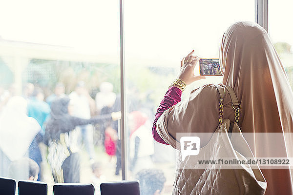 Rückansicht einer Frau  die durch ein Smartphone fotografiert  während sie am Fenster steht