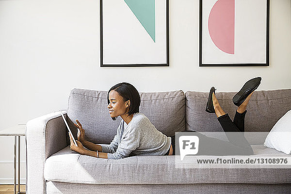 Seitenansicht einer Frau  die einen Tablet-Computer benutzt  während sie zu Hause auf dem Sofa liegt