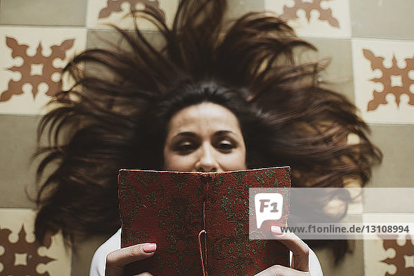 Hochwinkelansicht einer Frau  die ein Buch liest  während sie zu Hause auf dem Boden liegt