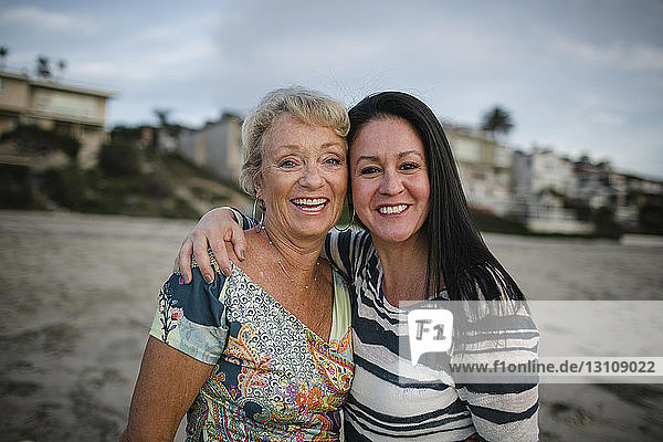 Portrait of smiling mother and daughter standing at beach against sky during sunset