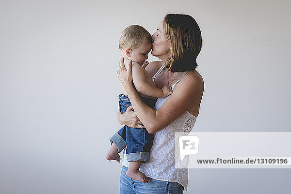 Mother kissing shirtless son while carrying him against white background