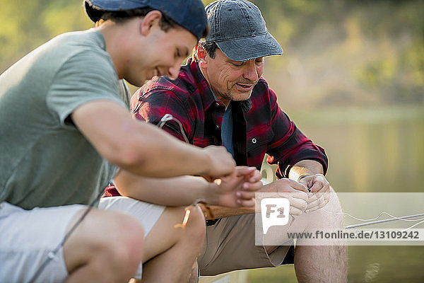 Male friends adjusting fishing rods while sitting at lakeshore