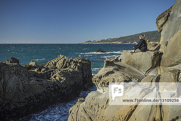 Side view of woman sitting on rocks at beach against clear sky