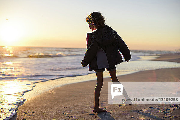 Girl in jacket looking at sea while standing at beach during sunset
