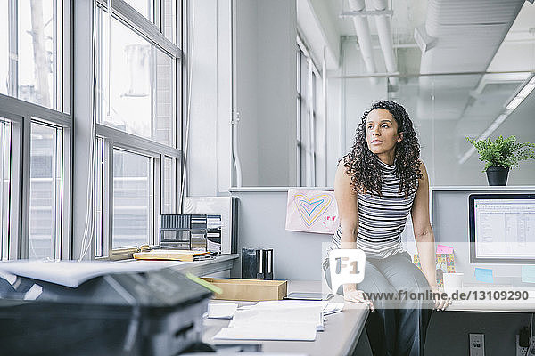 Businesswoman looking through window while sitting on desk in office