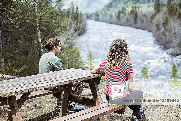 Rückansicht von Geschwistern  die auf den Fluss schauen  während sie auf dem Picknicktisch sitzen