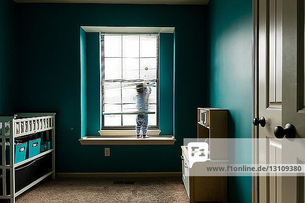 Rückansicht eines Jungen  der mit Jalousien spielt  während er zu Hause auf der Fensterbank steht