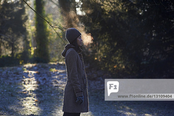 Seitenansicht einer Frau  die Atemdampf ausatmet  während sie im Winter im Park steht