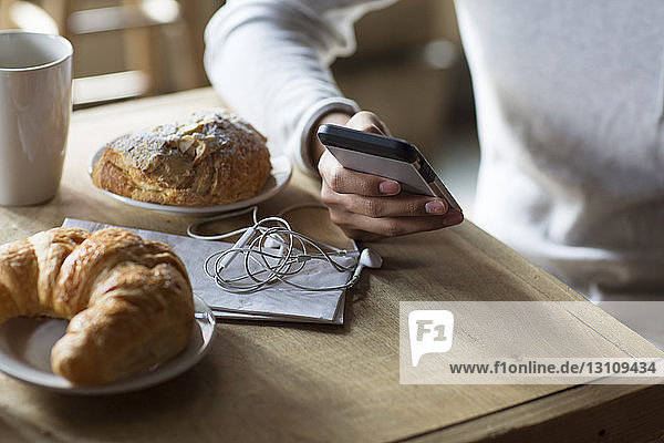 Cropped image of man holding mobile phone by croissants and coffee at table