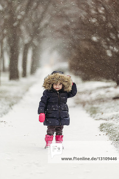 Portrait of cute girl wearing hooded jacket while standing on snowy footpath during snowfall