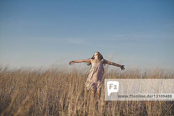 Glückliches Mädchen mit ausgestreckten Armen auf trockenem Grasfeld vor blauem Himmel stehend