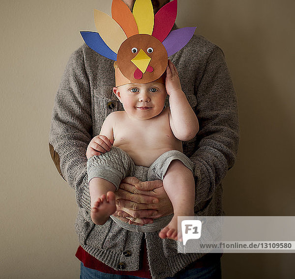 Portrait of cute shirtless son wearing colorful paper headdress while being carried by father against wall at home