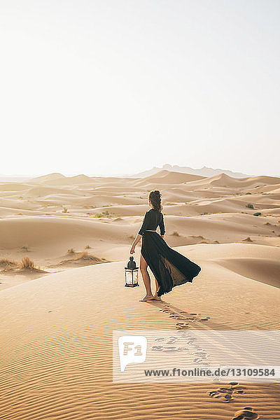 Seitenansicht einer Frau  die eine Laterne hält  während sie bei Sonnenuntergang in der Wüste Sahara vor klarem Himmel steht