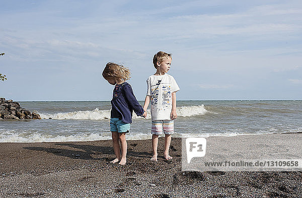 Süße Geschwister halten sich an sonnigen Tagen am Strand an der Hand  während sie am Strand gegen den Himmel stehen