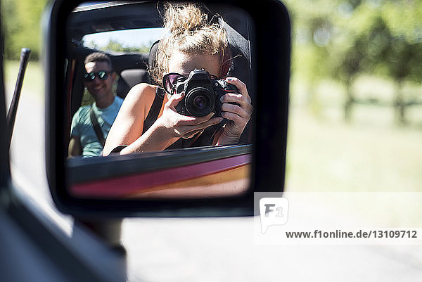 Frau fotografiert sich im Seitenspiegel reflektierend  während sie mit einem Freund im Geländewagen unterwegs ist