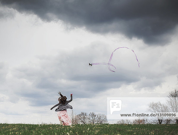 Rückansicht eines spielerischen Mädchens das einen Drachen gegen einen bewölkten Himmel im Park fliegen lässt