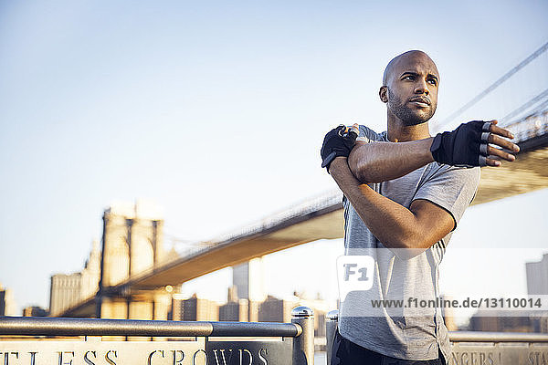 Entschlossener männlicher Athlet macht Dehnungsübungen auf der Promenade mit der Brooklyn Bridge im Hintergrund vor klarem Himmel