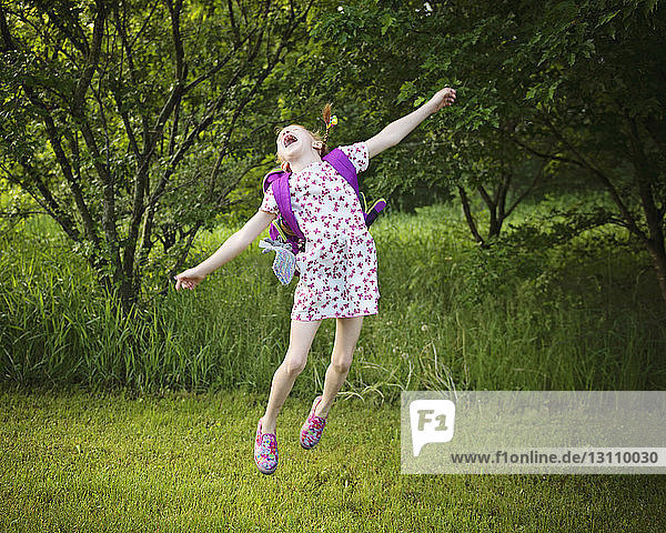 Cheerful girl with backpack jumping over grassy field at park