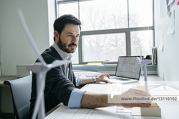 Geschäftsmann benutzt Laptop  während er das Modell einer Windkraftanlage auf dem Schreibtisch arrangiert