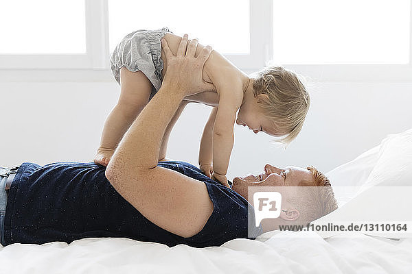 Side view of playful father lifting shirtless daughter while lying on bed at home
