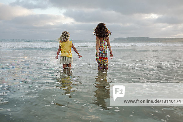 Rear view of sisters standing in sea against cloudy sky