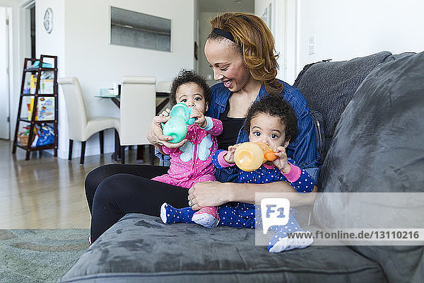 Happy mother holding daughters drinking milk while sitting on sofa at home