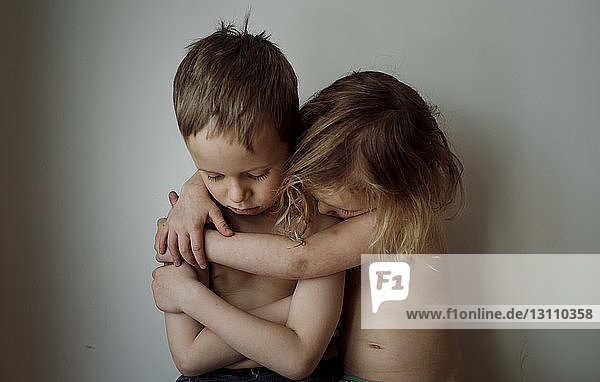 Schwester ohne Hemd umarmt traurigen Bruder zu Hause an der Wand