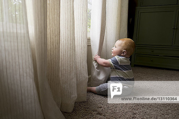 Seitenansicht eines kleinen Jungen  der mit Vorhängen spielt  während er zu Hause auf einem Teppich sitzt