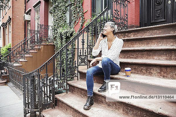 Happy woman talking on mobile phone while sitting on steps with coffee cup