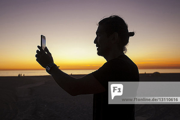 Seitenansicht eines Mannes  der mit einem Smartphone vor dramatischem Himmel am Strand bei Sonnenuntergang fotografiert