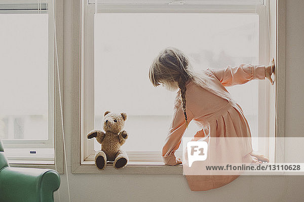 Seitenansicht eines Mädchens  das einen Teddybär anschaut  während es zu Hause auf dem Fensterbrett kniet