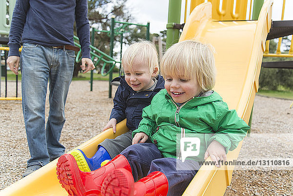 Low section of father standing by sons playing on slide at playground