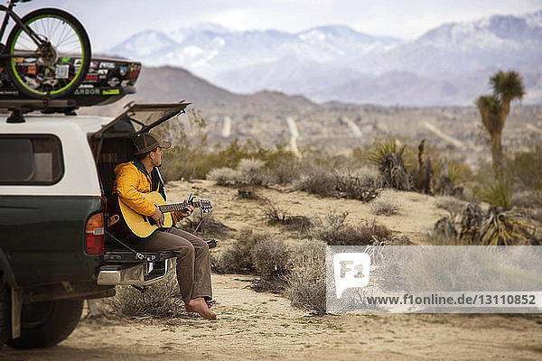 Mann spielt Gitarre  während er auf dem Kofferraum eines Autos sitzt