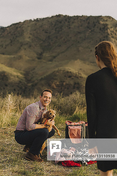 Freund trägt Hund  während er seine Freundin auf dem Feld gegen die Berge ansieht