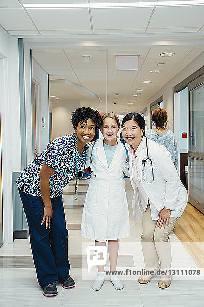 Full length portrait of cheerful female doctors with girl wearing lab coat in hospital corridor