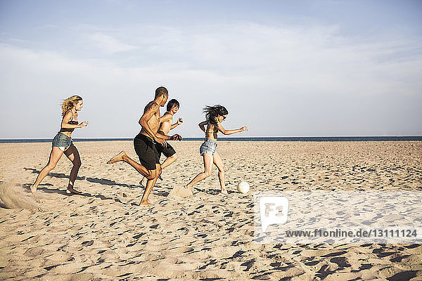 Friends playing soccer on sand at beach against sky