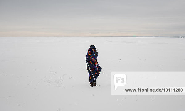 Rückansicht einer in eine Decke gehüllten Frau  die auf einer schneebedeckten Landschaft vor dem Himmel steht