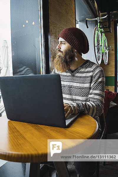 Hipster-Mann schaut durchs Fenster  während er seinen Laptop-Computer auf dem Tisch im Café benutzt