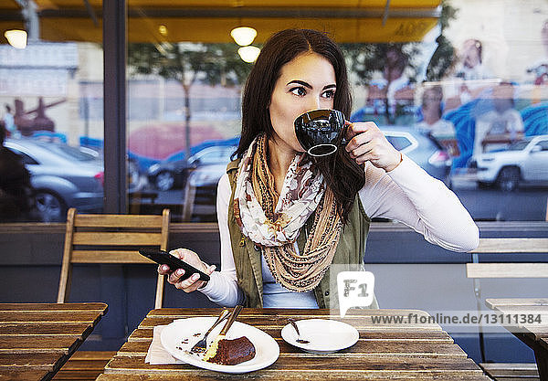 Frau hält Mobiltelefon in der Hand und genießt ein Getränk im Café