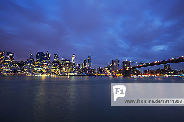Blick auf die beleuchtete Skyline und die Brooklyn Bridge bei bewölktem Himmel in der Nacht