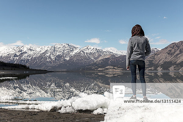 Frau in voller Länge auf Schnee am Fluss stehend  während sie gegen Berge und Himmel blickt
