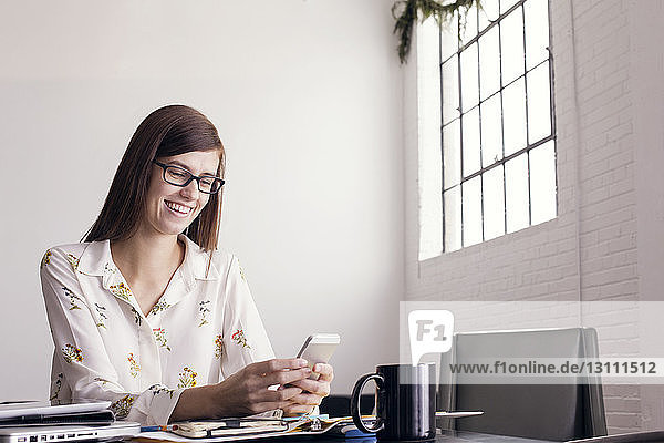 Lächelnde Geschäftsfrau benutzt Telefon  während sie am Tisch sitzt