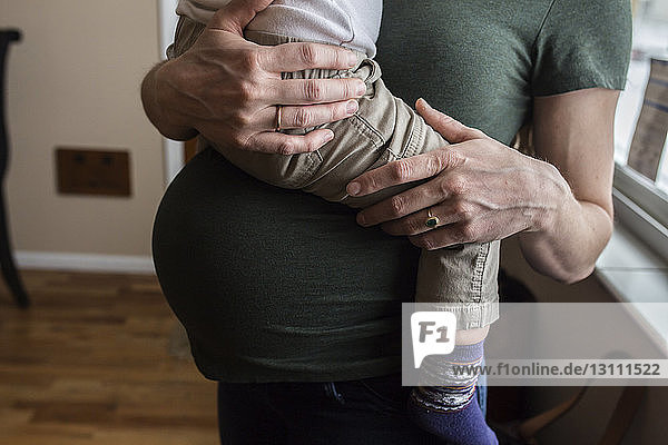 Mittelsektion einer schwangeren Mutter  die ihren Sohn zu Hause gegen das Fenster trägt