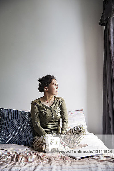Nachdenkliche Frau sitzt zu Hause mit Laptop auf dem Bett