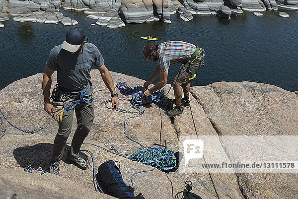 High angle view of friends tying climbing ropes on rock formation over lake during sunny day
