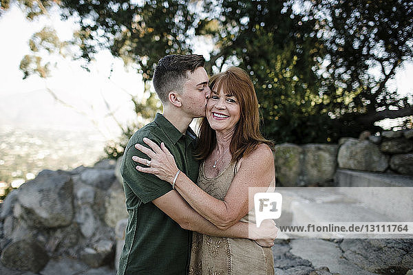 Seitenansicht eines glücklichen Sohnes  der seine Mutter küsst  während er im Park an Bäumen steht