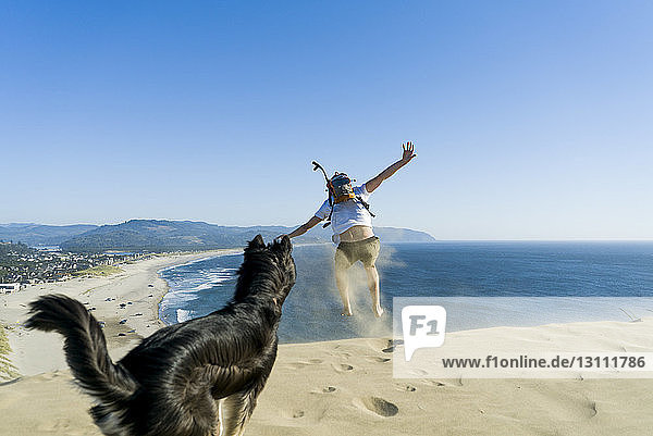Verspielter Mann springt  während der Hund am Strand vor klarem blauen Himmel steht
