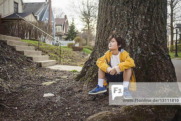 Thoughtful boy looking away while sitting by tree trunk