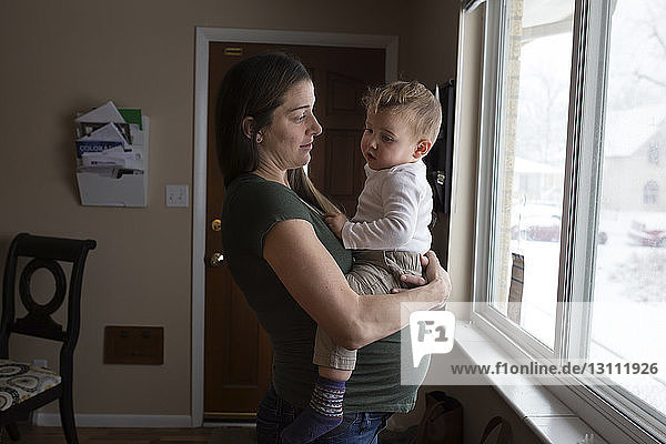 Seitenansicht der schwangeren Mutter  die den Sohn ansieht  während sie ihn gegen das Fenster trägt